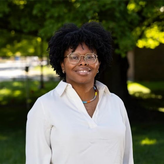 A person wearing glasses and a white shirt stands outside in front of green trees, smiling at the camera.