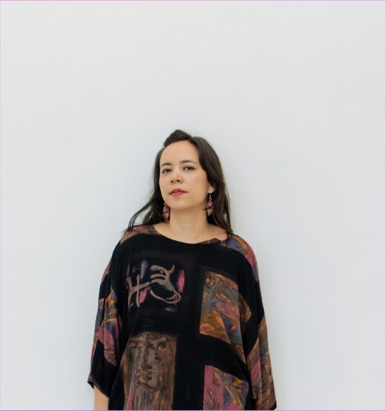 A woman with long brown hair stands against a plain white wall, wearing a dark, patterned top and red earrings.