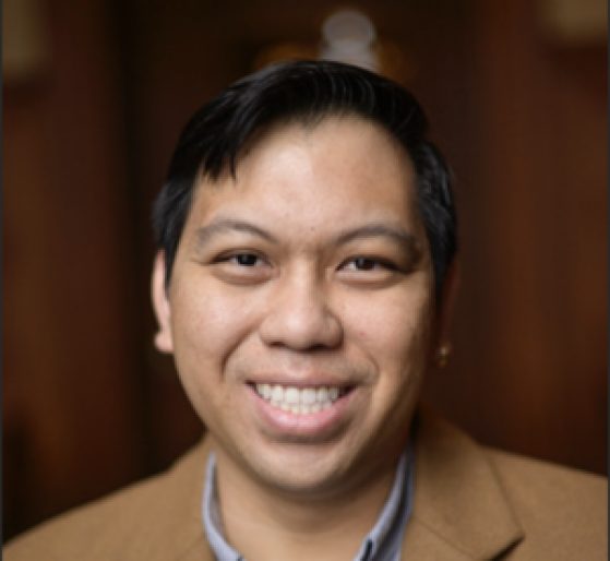 A person with short black hair and a tan blazer smiles at the camera against a blurred brown background.