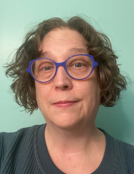 A person with short curly hair, wearing blue glasses and a textured blue top, poses in front of a light teal background.