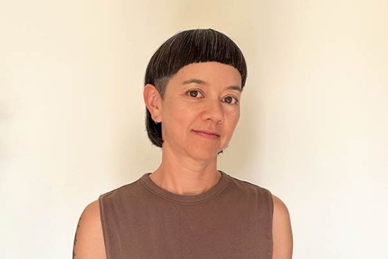 Person with short, dark hair and bangs wearing a sleeveless brown top stands in front of a plain, light-colored background.