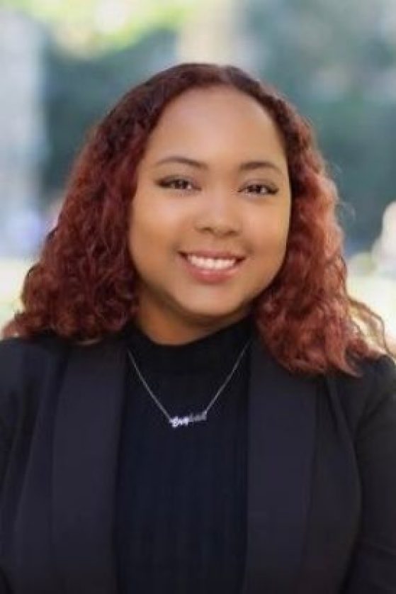 A person with curly, shoulder-length reddish hair is smiling, wearing a black top, black blazer, and a necklace, with a blurred outdoor background.