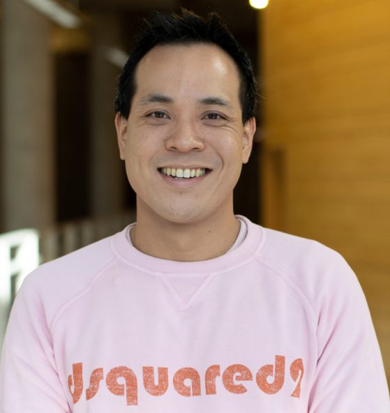 A person with short black hair is smiling, wearing a light pink sweatshirt that says Dsquared2, standing indoors with a blurred background.