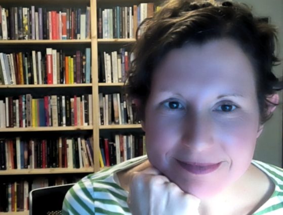 Person with short, curly hair wearing a green and white striped shirt, sitting in front of a bookshelf filled with books, smiling at the camera.