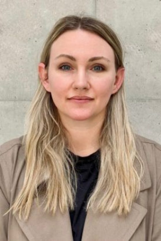 A woman with long blonde hair and a neutral expression stands in front of a light gray concrete wall, wearing a beige coat over a black top.