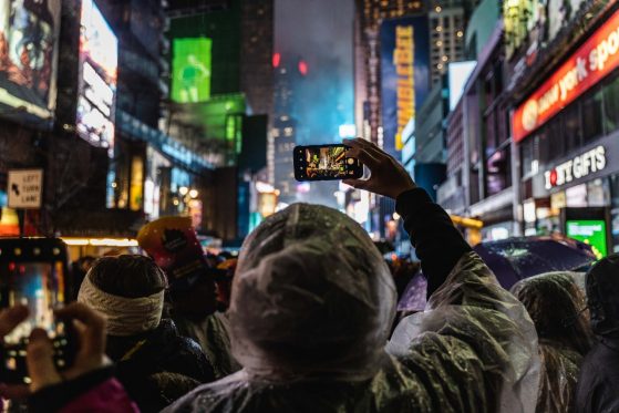 A person taking a picture with their phone is in focus with the blurry city lights behind it. 