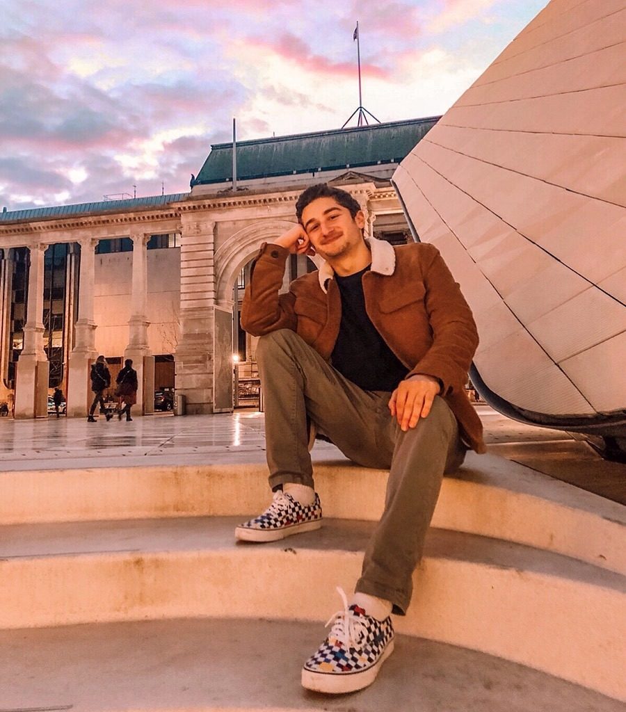 Adam poses seated on some steps in London. There is an older building behind him and the sky is vibrant with color.