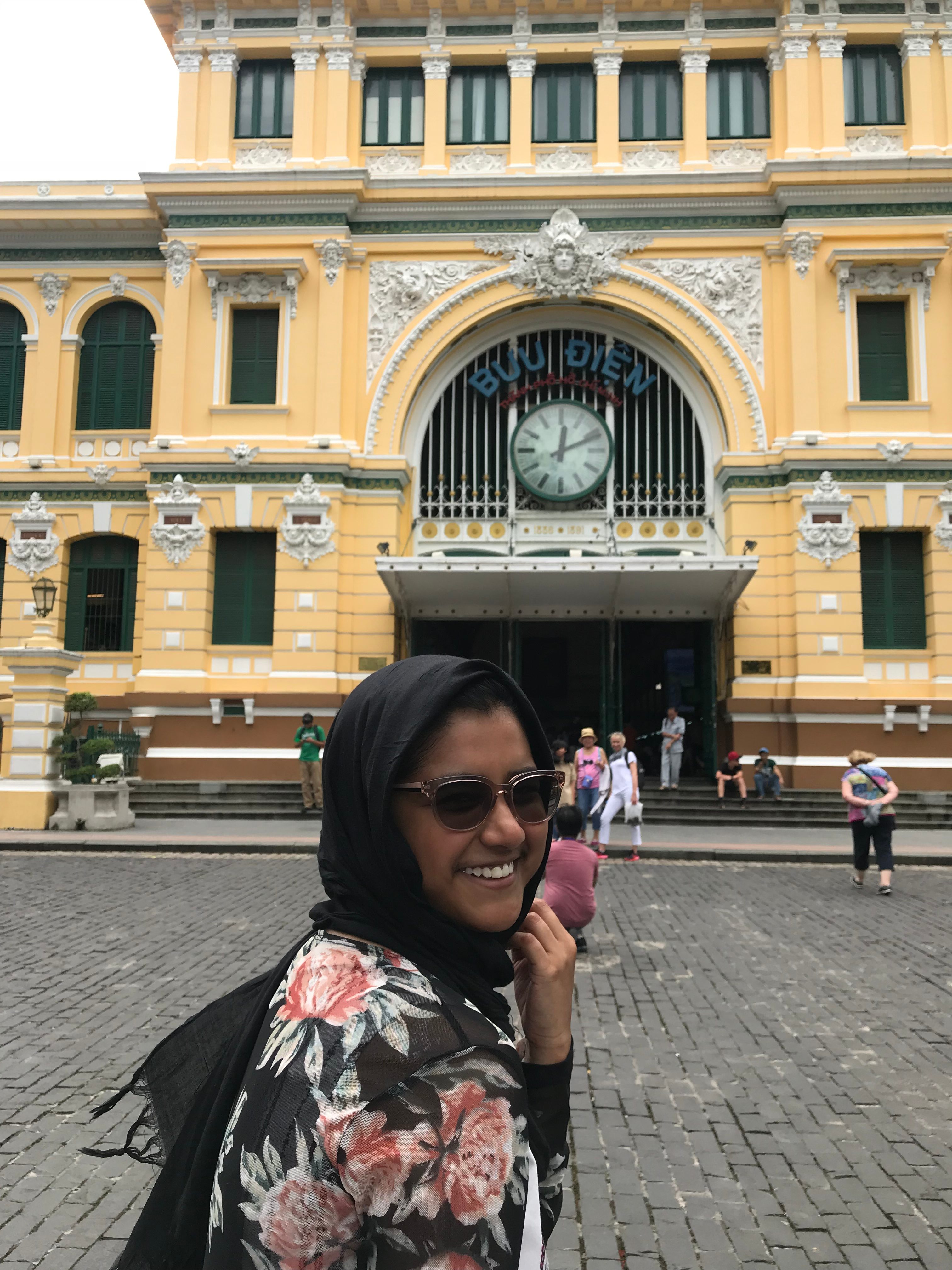 Tazia pauses while walking down a street to pose for a picture in front of a yellow brick building with white accents. 