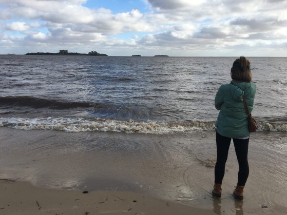 A student stands on the beach staring at the ocean. They are wearing a teal light-coat and watching the waves.