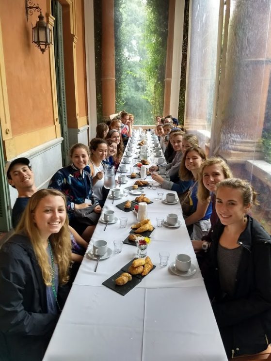 Students lean in eagerly around a long, rectangular table, their eyes sparkling as they gaze at the camera.