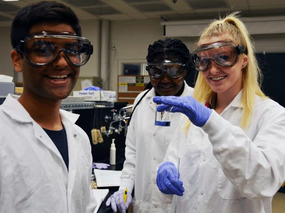 three students in lab coats, gloves and googles look at camera. one young woman holds up beaker with blue fluid in it