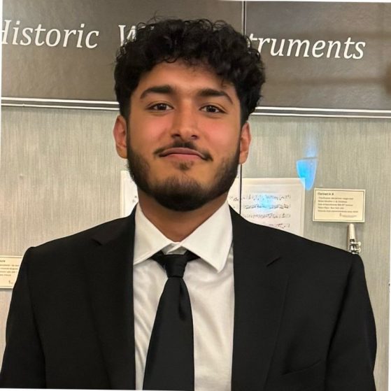 Aryan Singh smiling in a black suit and tie, before a musical exhibit.