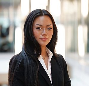 April Xiao standing before a well-lit but blurred building interior background in a black business suit.