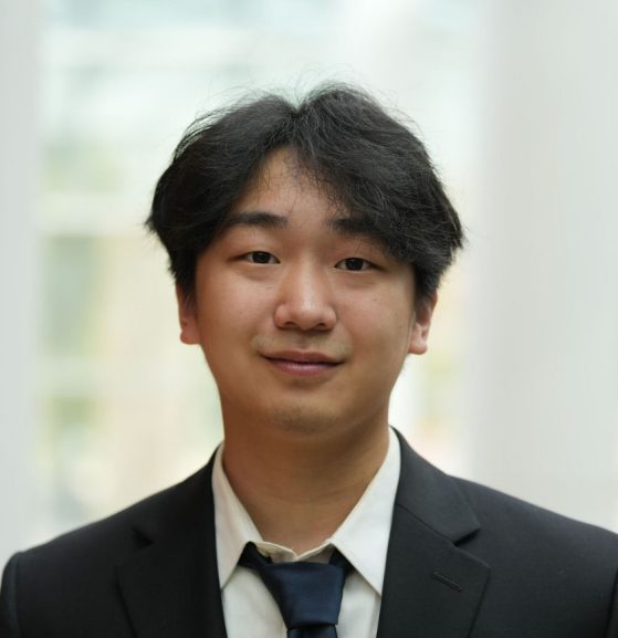 Jihan Park smiling in a black suit and dark blue tie with a well-lit interior softly blurred in the background