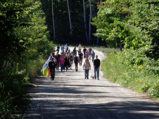 A group of about 30 people walking Indian Road to the second home of the Burt Lake Band, one carrying their flag, another walking a dog. The winding road is surrounded by dense woods.