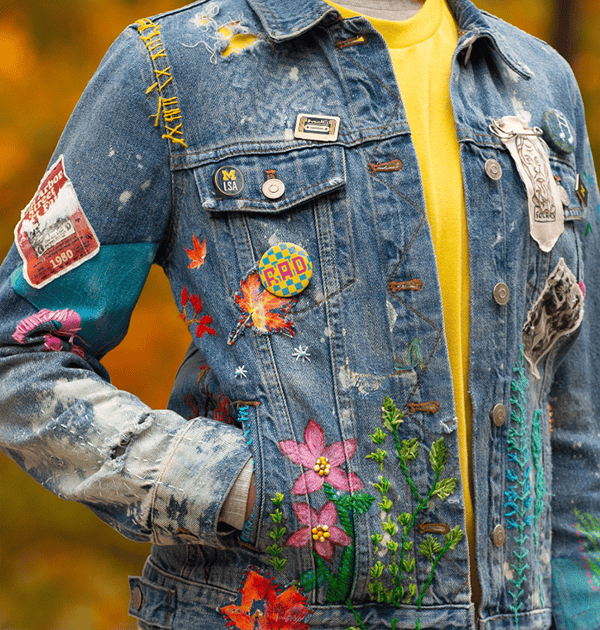 A photo of a person wearing a denim jacket. The jacket includes embroidered flowers, a variety of pins and buttons, paint, a handmade fabric piece with a drawing of Socrates, and faded spots that suggest many years of wear.