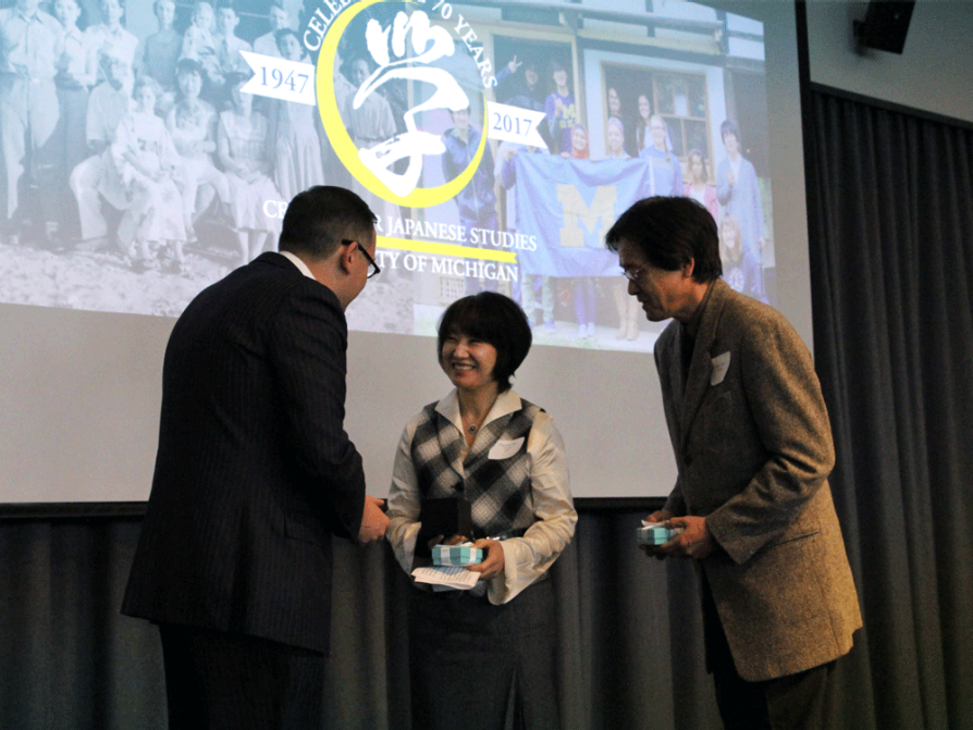 Mayumi and Masao Oka are facing LSA Dean Andrew Martin onstage. They are each holding light blue boxes tied with ribbon. The Center for Japanese Studies logo is on the screen behind them.