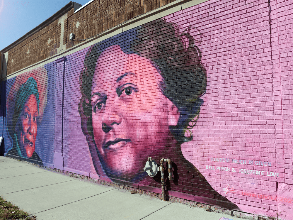 Color photograph of the mural component of To Whom Much Is Given. In vibrant shades of pink with purple and blue transitions, a mural by Ijania Cortez lives in permanence. On the left, a likeness of Malika Pryor’s grandmother, Dell Pryor, looks out over West Forest Avenue. On the right, a likeness of Josephine Love faces her former contemporary and looks upward in what could be interpreted as contemplation. Behind Love, bolded white lettering solidifies the phrase “To Whom Much Is Given” and names the two Detroit arts visionaries.