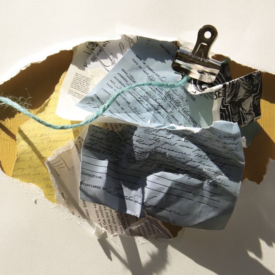 Several fragments of crumpled paper resting in a larger paper fragment. There is a binder clip and a strand of yarn. The writing is not legible.