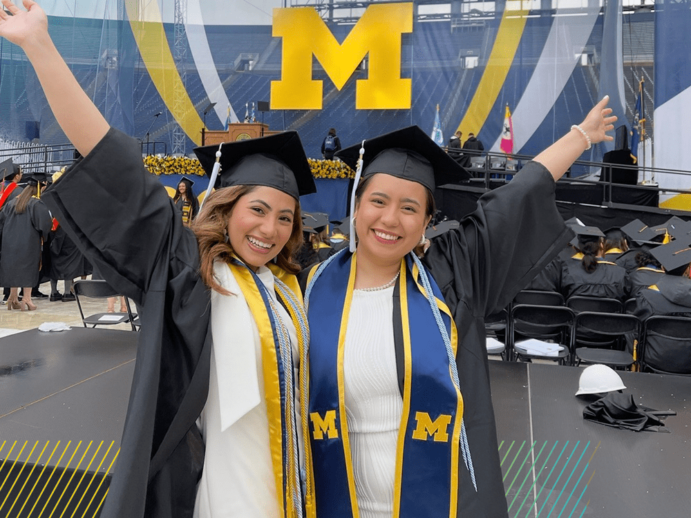 Two U-M graduates in caps and gowns celebrate on graduation day. They each hold one arm in the air. A maize Block M hangs behind them.