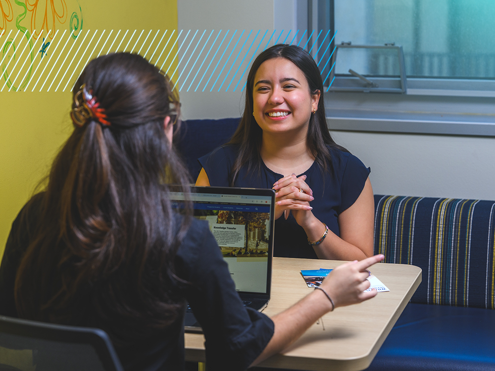 Two people sit at a table, one with her back to the camera and one, smiling, facing the camera.