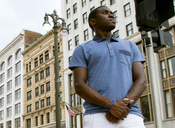 A photograph of a young man wearing a light blue short-sleeved shirt. He's standing on a city street, near a crosswalk.