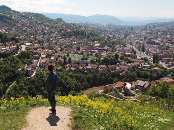 Hunter Zhao standing atop a hill and looking down at the Bijela Tabija ("White Castle:) historical district in Sarajevo, a town nestled into a valley. Mountains are visible in the distance. 