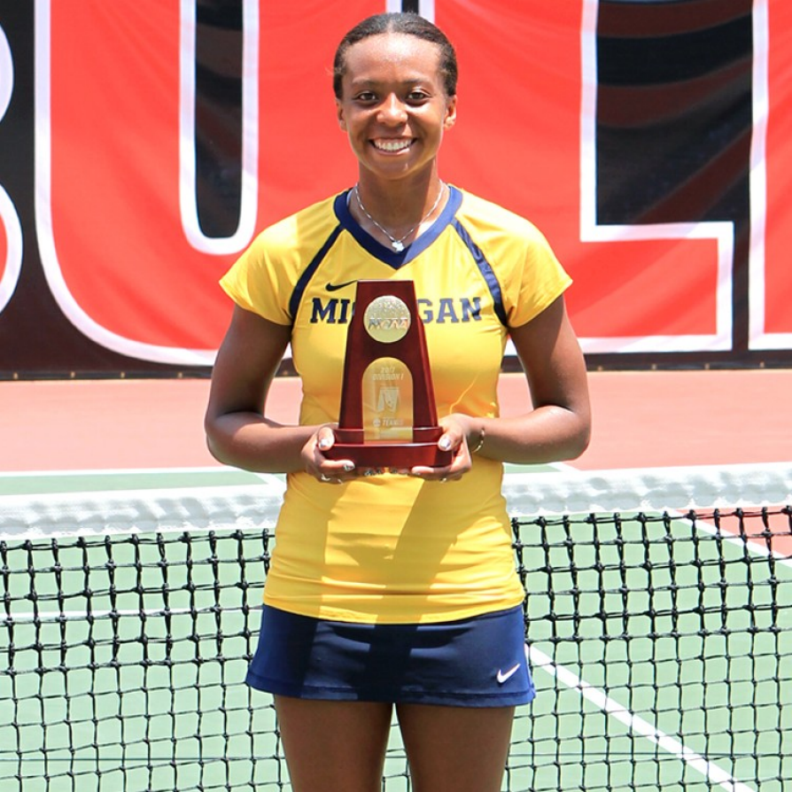 Brienne Minor stands in front of the net on a tennis court wearing her gold U-M tennis team uniform. She is holding a trophy .