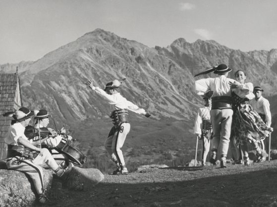 A black and white photo of people dancing on a ridge with a range of the mountains behind them at a distance. The people are wearing costumes: the men wear feathered caps and hold poles; the woman wears a dress with a long full skirt. At the left edge of the photograph, one man is playing a cello and the other is playing a violin.