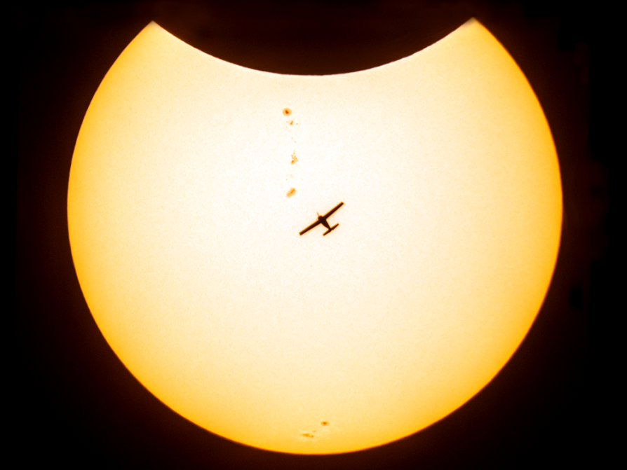 a photograph of a yellow sphere with the edge of a dark sphere floating into its top. A silhouette of an airplane is angled toward the top left. There are a few small spots on the yellow surface.