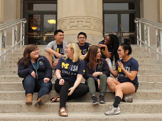 Seven Kessler Presidential Scholars sitting on the front steps of Angell Hall in a group talking to each other.