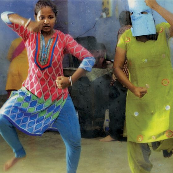 Two women dancing next to each other. One woman wears a red tunic and stands on one foot, arms akimbo. The other woman is wearing a blue mask over her face.