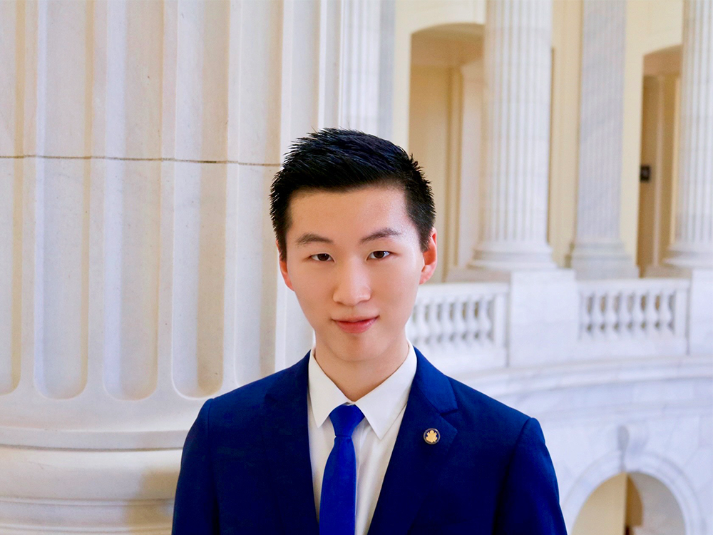 Shuyan Tang, dressed in a dark blue blazer, white dress shirt and blue tie, poses inside of a building.