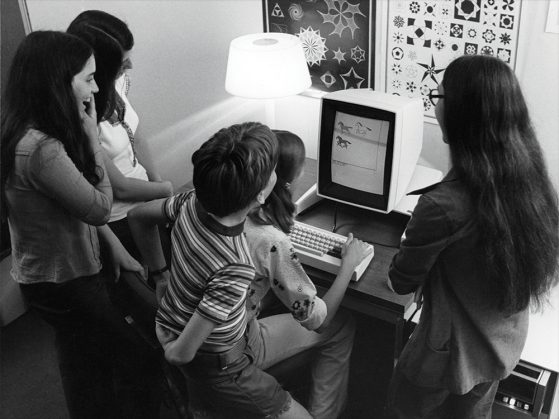 A black and white photograph of five kids gathered around an old computer screen. The screen displays three horses