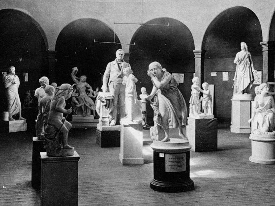 A black and white photograph of a collection of sculptures in a room with arches and mottled light.