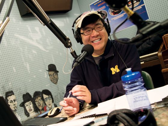 Robert Yoon behind the mic at WCBN. He's wearing headphones, a baseball hat, and a U-M fleece, and he's smiling at someone off camera.