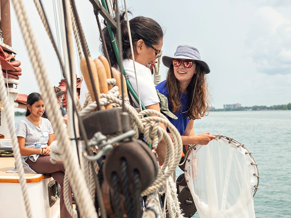 An educator onboard the Inland Seas schooner in the Detroit River teaches a faculty member how to catch plankton with a net. The instructor wears a hat and sunglasses and smiles toward the faculty member. Ropes and pullies are in the foreground.