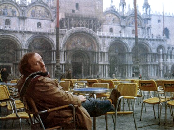 Joseph Brodsky in a Russian square, sitting at an empty outdoor cafe with yellow chairs. He is smoking a cigarette and he has his feet propped up on a chair.