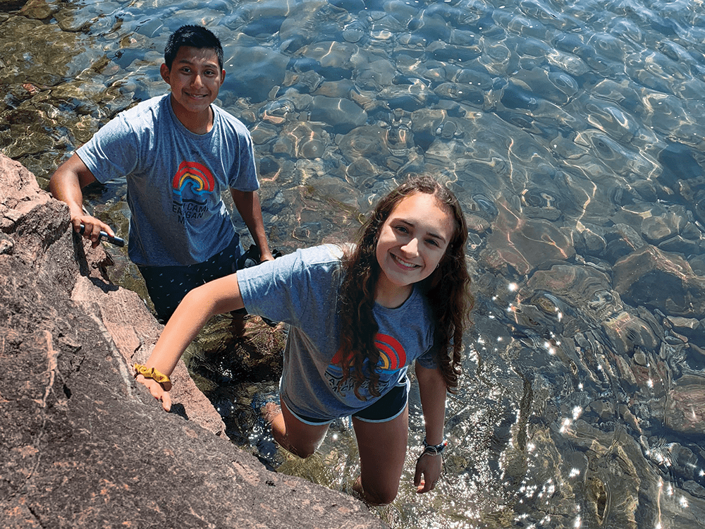 Two students stand near a rock in shallow water looking up