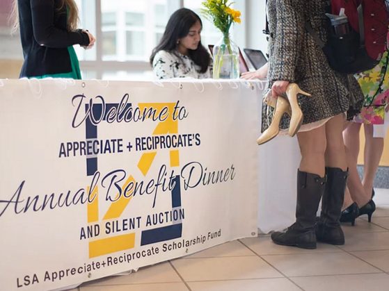 A table with a banner for the Appreciate-Reciprocate scholarship dinner. One student sits behind the table, and two women stand in front of it seen from the waist down. One of the women is carrying pumps and wearing winter boots.