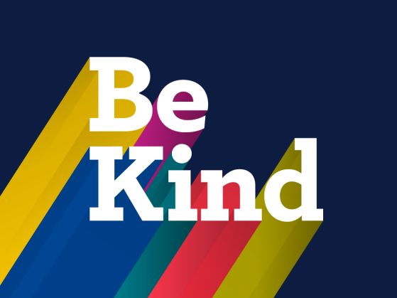 The image is an illustration of the words "Be Kind" in a large, white font with a colorful shadow effect under it from the bottom left corner of the screen. The colors of the shadow are yellow, blue, purple, green, red, and yellow from left to right.