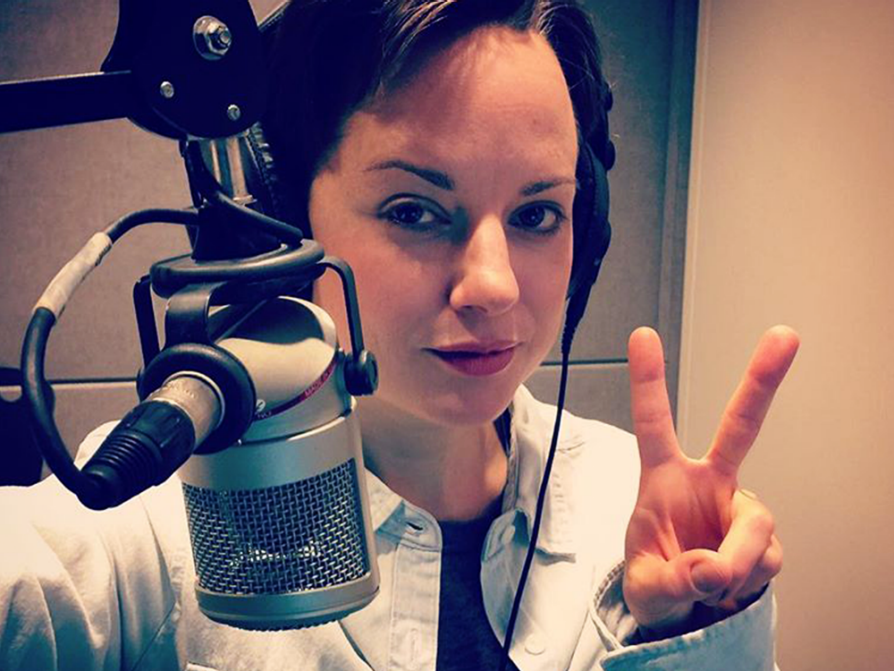 Jones-Rooy stands behind a broadcasting microphone holding the index and middle fingers up as a peace sign. She's wearing headphones.