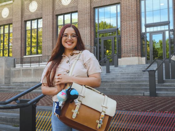 A woman wearing glasses and holding a leather messenger bag smiles directly at the camera.
