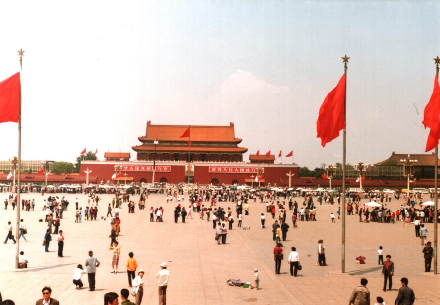 A photograph of Tiananmen Square taken in 1988. There are three red flags in the foreground and small groups of people standing or talking together in the square.