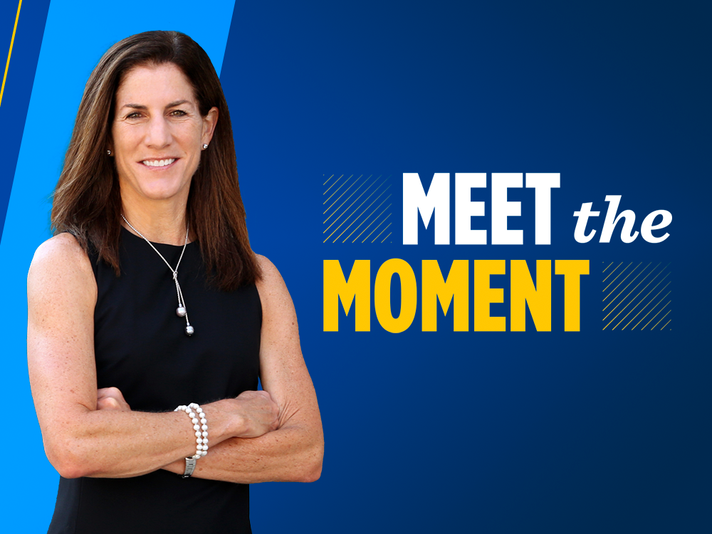 Dean Anne Curzan stands with arms crossed in front of a blue banner that reads “Meet the Moment.”