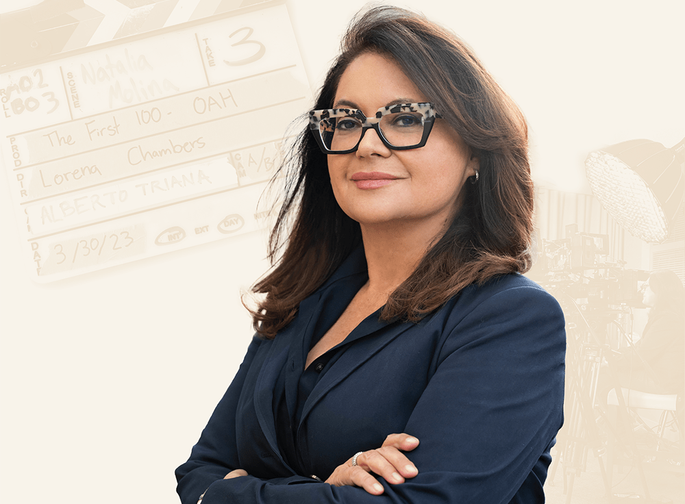 Lorena Chambers stands with her arms crossed in a blue blazer. She also wears a leopard patterned and black rimmed pair of glasses and gazes at the camera. In the background is of a movie clapperboard. The clapperboard has readouts about the production of Chambers’s project The First 100. In the background, a film crew with lighting and cameras works within a studio production space.