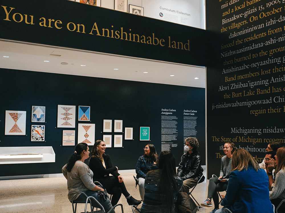 Students sit in a circle around artist Andrea Carlson as she speaks. Those pictured are in UMMA’s vertical gallery space. The Future Cache exhibit surrounds them, paintings and text on the wall. The text, which also tells the story of the Burt Lake Band, is white and yellow on a black background. Above the group, the largest words say ”You Are on Anishinaabe Land.”