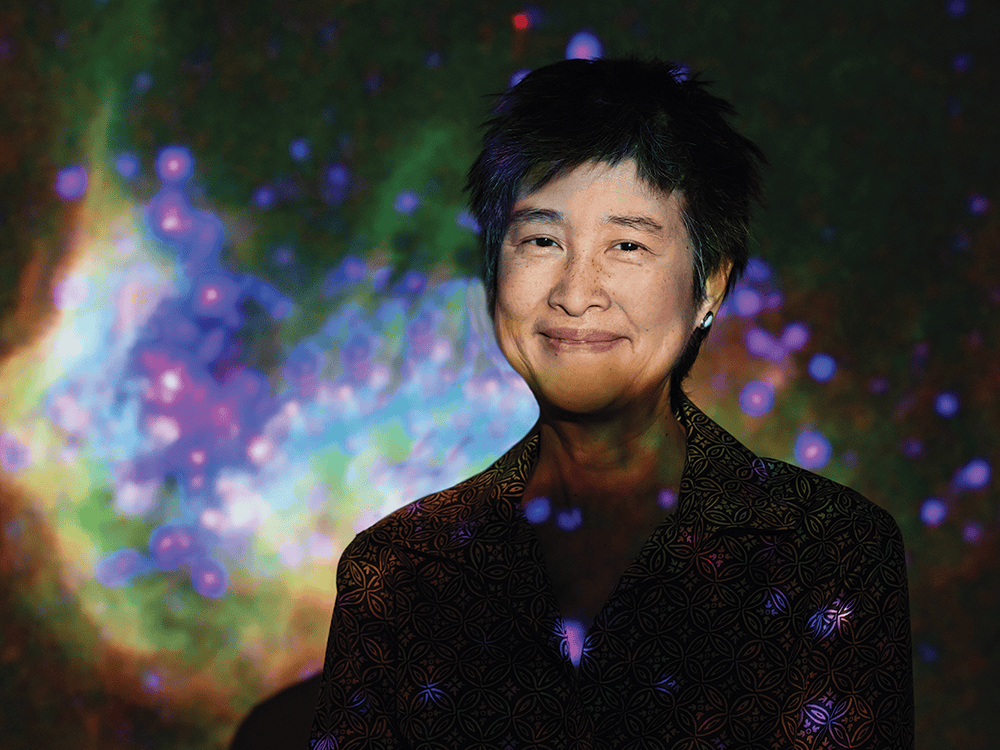 A head-and-shoulders photo of professor Sally Oey shows her facing the camera and smiling gently. Oey, wearing a gray geometric-pattern blazer, is angled slightly toward the left side of the image. The background of the image is a picture of a galaxy, with bright red, purple, blue, and green colors.