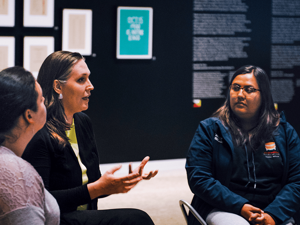 Artist Andrea Carlson speaks with two students during a talk at the University of Michigan Museum of Art. They are all seated. In the background is a portion of the exhibit of Carlson’s works, called Future Cache, which explores the Burt Lake Burn-Out, the forced relocation of the Burt Lake Band of Ottawa and Chippewa Indians.