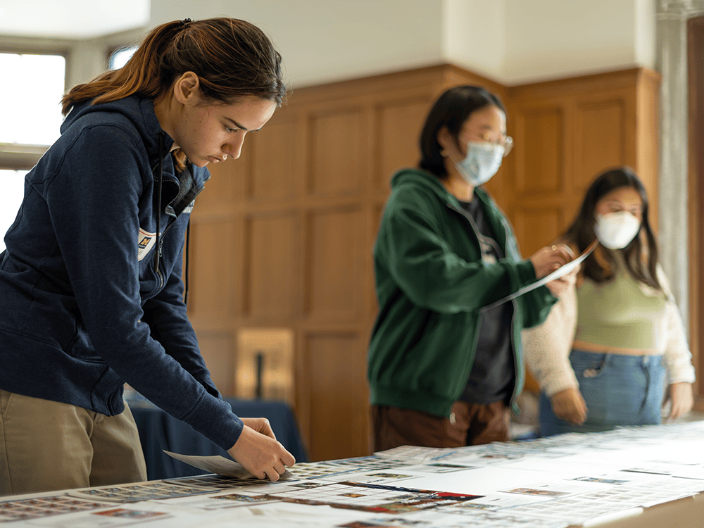 Three female undergraduate students review materials on a table. Two of the students wear masks and one does not. The students all look focused on the papers that are strewn on the table.